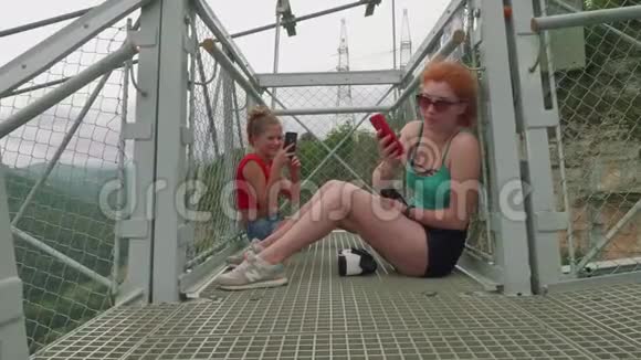 两个白种人金发碧眼的人坐在山上高高的秋千桥上在智能手机上拍照视频的预览图