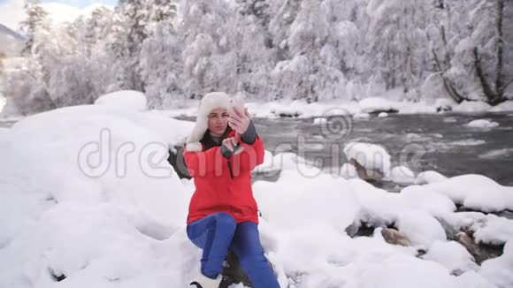 穿着红色夹克和毛皮帽的女孩在山上肆虐的冬季河流旁边的智能手机上自拍视频的预览图