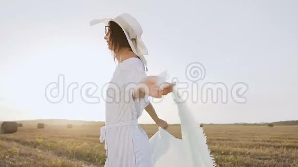 一个穿着长裙戴着墨镜戴着围巾的大帽子的迷人女孩在田野里旋转的肖像视频的预览图