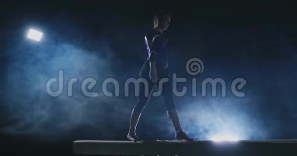 职业体操运动员女孩在平衡木上缓慢地旋转一跳健身房里的蓝色烟雾视频的预览图