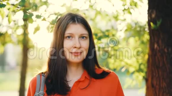 一位棕色头发的美丽女孩的特写肖像看着镜头在背景中带着夏日公园微笑视频的预览图