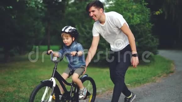 带着细心的父亲在公园里骑自行车笑孩子骑自行车的慢动作年轻快乐视频的预览图