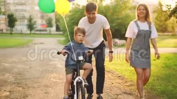 可爱的父母在教儿子可爱的男孩在城市公园骑自行车孩子在骑自行车他的父亲抱着他视频的预览图