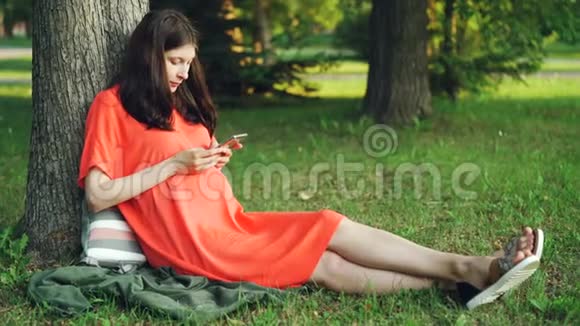 迷人的孕妇穿着休闲服装正在使用智能手机坐在公园树下的草地上怀孕人视频的预览图