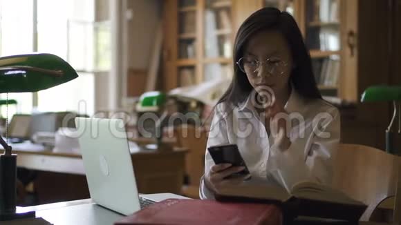 年轻的生物学家正在用智能手机坐在桌子上图书馆里有笔记本电脑视频的预览图