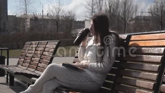 公园里一个穿着运动服的漂亮女孩坐在长凳上看书喝着热杯子里的咖啡大笑视频的预览图