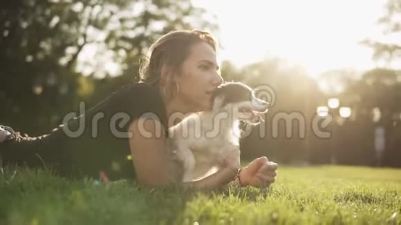 侧视镜头一名穿着黑色T恤的白种人年轻女子和她的小狗躺在草地上看着视频的预览图