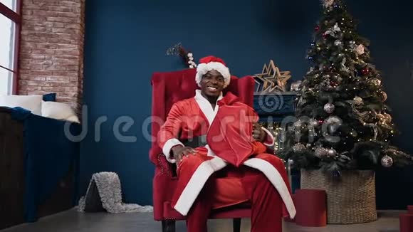 漂亮的非洲男人穿着圣诞老人服装和帽子拿着礼物坐在圣诞树附近的椅子上视频的预览图
