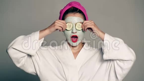 一个女孩脸上戴着面具头上戴着毛巾用灰色背景上的黄瓜闭上眼睛妇女视频的预览图