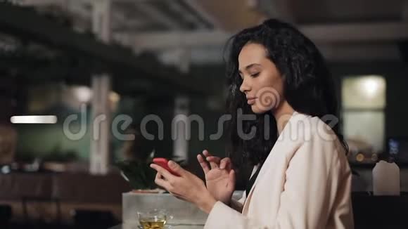 漂亮的年轻女商人在舒适的咖啡馆里使用智能手机女孩微笑着在网上和朋友聊天视频的预览图