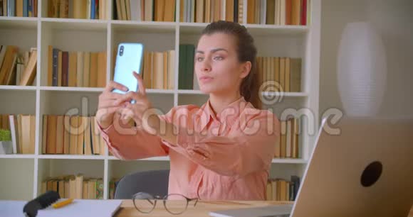年轻漂亮的女商人坐在笔记本电脑前的手机上自拍的特写视频的预览图