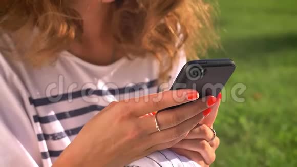 年轻的白种人黑发女人坐在草地上的公园里用智能手机滚动打字背景是大学视频的预览图