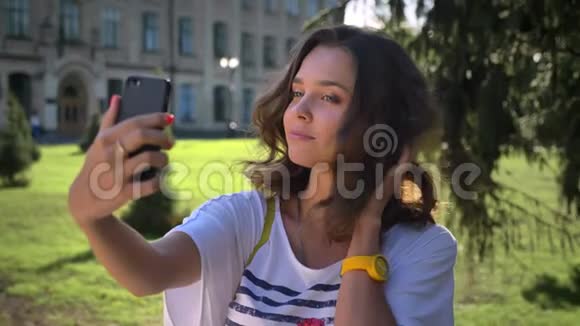 一个年轻的白人女孩站在公园里用智能手机自拍在大学里自拍的肖像视频的预览图
