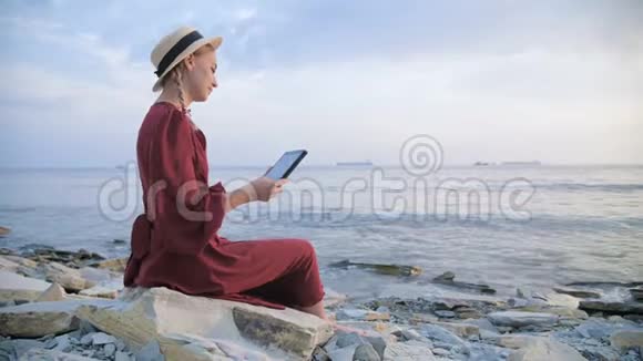 晚上一个穿着夏红色连衣裙和草帽的漂亮姑娘坐在海边的一块石头上看着视频的预览图
