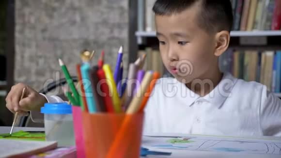 年轻的亚洲小男孩在纸上画画坐在桌子后面孩子做作业书架背景视频的预览图
