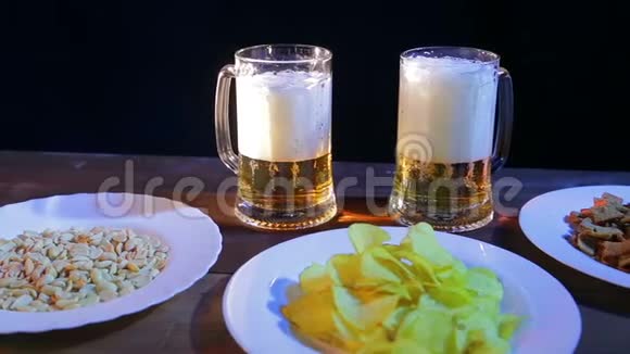 在黑色背景的酒吧里的木桌上放着两个起泡啤酒的杯子在盘子附近有薯片和坚果视频的预览图