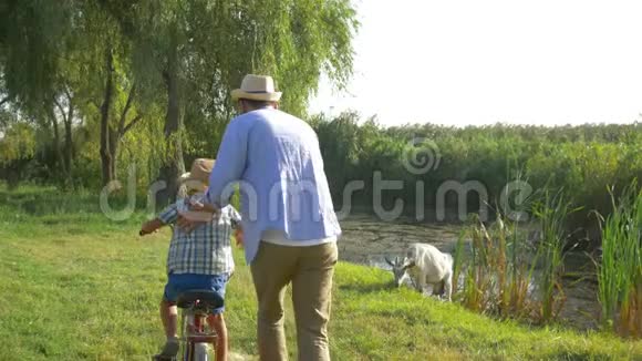 小男孩在爸爸的帮助下学习骑自行车背景是户外自然和小河视频的预览图