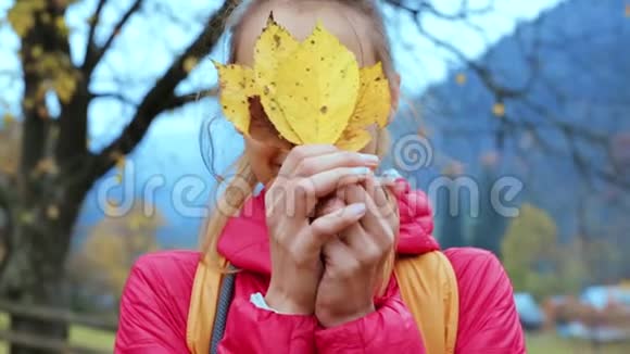 穿着红色夹克的漂亮微笑的女人站在户外手里拿着几片干黄的叶子和她们一起玩视频的预览图