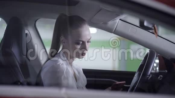 穿着白衬衫的可爱女性坐在车里拿着方向盘在车库旁边视频的预览图