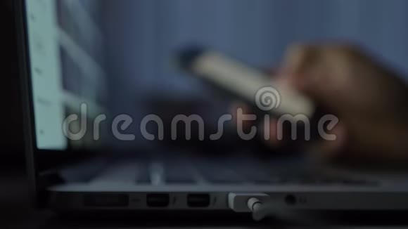 模糊的特写镜头手在智能手机前笔记本电脑在黑暗的背景下打字视频的预览图
