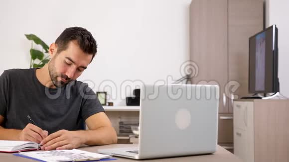 在家穿休闲服装的商人办公桌上放着笔记本电脑在笔记本上记笔记视频的预览图