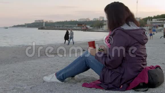 4一位穿春衣的漂亮女士端着一杯热饮料坐在海滩上用她的电话视频的预览图