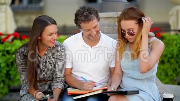 在阳光明媚的城市公园里坐在长凳上准备考试的三个朋友的前景视频的预览图
