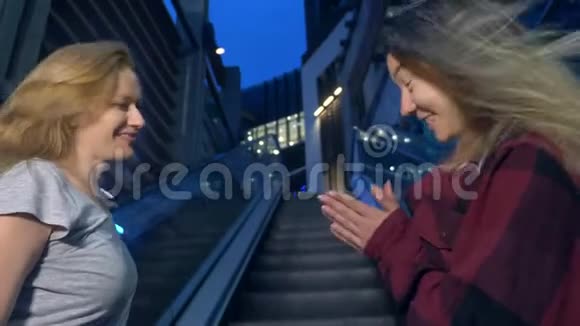 两个女孩亚洲人和高加索人晚上在现代城市的街道上聊天站在自动扶梯上视频的预览图