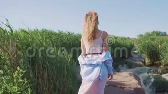 一个年轻的女孩穿着粉红色的裙子和蓝色的衬衫站在河的岩石海岸上背对着镜头视频的预览图