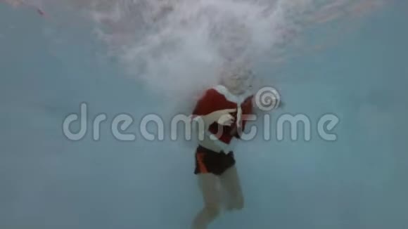 一个打扮成圣诞老人的小男孩在泳池里的喷水中游泳他的眼睛睁着微笑着看着摄像机视频的预览图