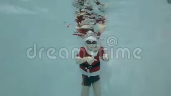 一个穿着西装戴着红色圣诞帽的快乐小男孩被淹没在游泳池里男孩在游泳微笑着看着视频的预览图
