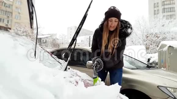 美丽微笑的年轻女人用刷子把雪从她的汽车上移走的4K镜头司机清洗车视频的预览图