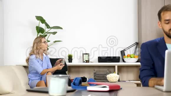 女人在控制台上玩电子游戏而她的男朋友在同一个客厅里电脑上工作视频的预览图