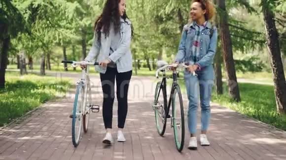 运动的年轻女人骑着自行车在公园里散步聊天欣赏美丽的杉树和温暖的春天他们视频的预览图