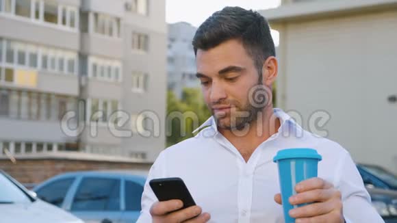 帅哥在他的智能手机上打字在办公楼附近的停车场上喝美味的咖啡看上去视频的预览图