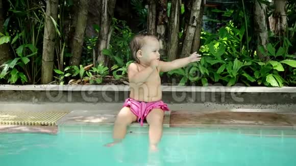 坐在泳池边的婴儿女孩坐在游泳池旁边用手把它拿出来慢动作视频的预览图