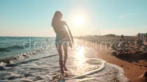 美丽苗条的女孩走在海滩上拿着冲浪板傍晚日落时跑步穿着泳装视频的预览图