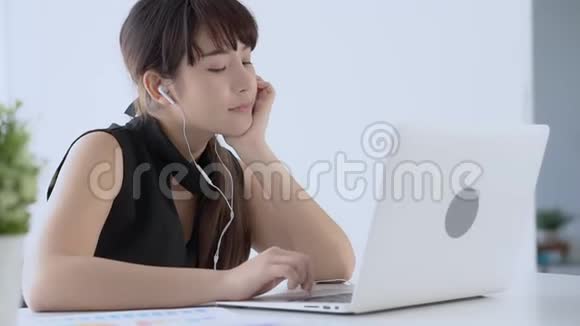 美丽的年轻亚洲女人微笑着坐在办公室里用手提电脑听带耳机的音乐放松视频的预览图