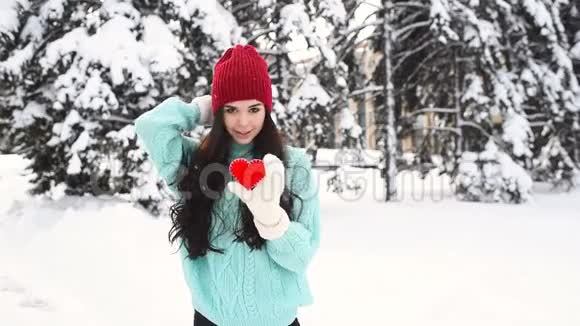 年轻漂亮的女孩穿着一件温暖的蓝色毛衣戴着手套背景是白雪覆盖的树木抱着一颗红色的心视频的预览图