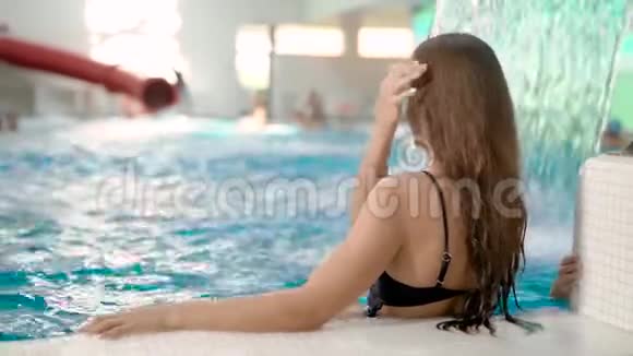 从一个漂亮的黑发女人坐在室内游泳池的背后观看视频的预览图