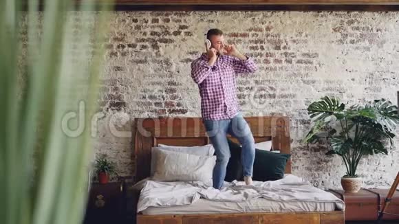 小胡子男人在双人床上跳舞用耳机听音乐的慢动作现代阁楼式公寓视频的预览图