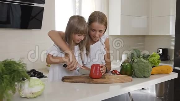 两个穿着白色连衣裙的可爱姐妹在厨房做饭女孩们切红辣椒做沙拉姐姐教老师视频的预览图