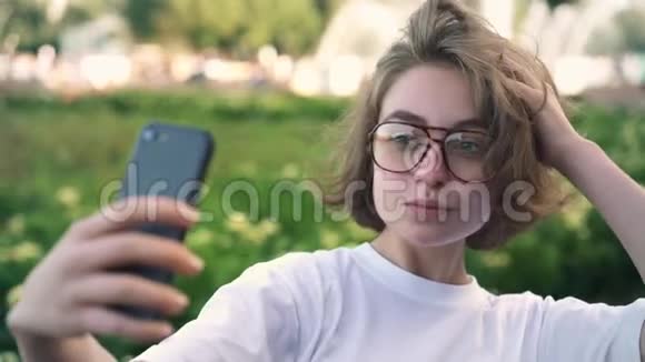 可爱的年轻女孩戴着眼镜在夏天的公园里拍自拍或浏览视频的预览图