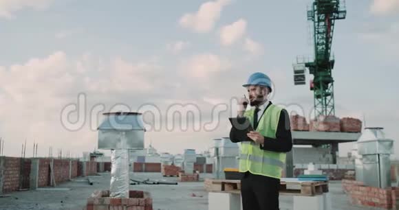 年轻的商人穿着一套安全设备在楼顶上和一些工人用收音机说话视频的预览图