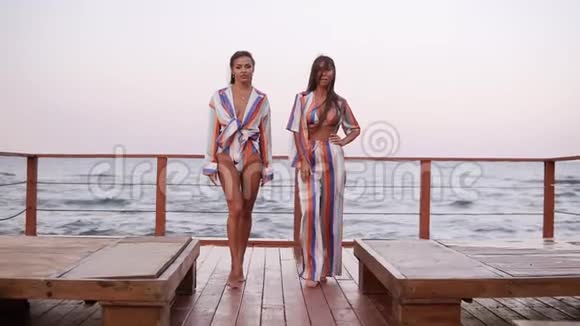 两个迷人的女孩在夏天穿着相同颜色的时尚衣服在感官上行走的慢动作镜头海洋区视频的预览图