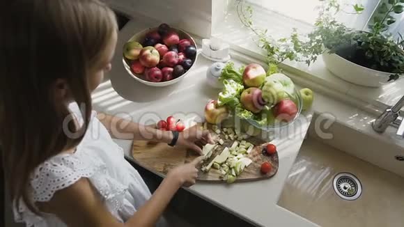 上景漂亮的金发女孩穿着白色连衣裙正在厨房准备水果沙拉女孩把一个苹果切成薄片视频的预览图