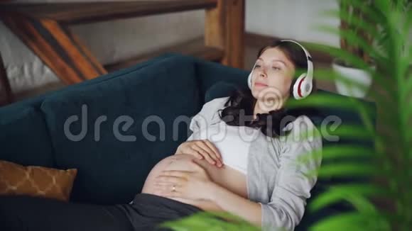 穿着漂亮的孕妇躺在沙发上抚摸着肚子用耳机听音乐视频的预览图