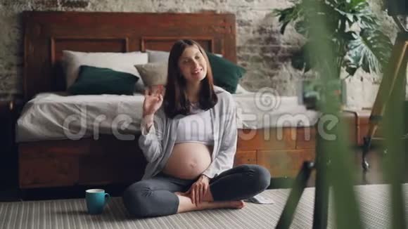 美丽的年轻女性博客写手正在为她的Vlog录制关于怀孕的视频她坐在地板上聊天聊天视频的预览图