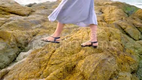特写镜头女人穿着凉鞋腿和一条长长的灰色裙子在低潮的岩石海岸上视频的预览图