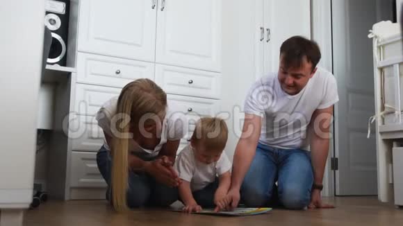 一家三口穿着白色T恤衫和蓝色牛仔裤坐在卧室在卧室的地板上和男孩玩视频的预览图
